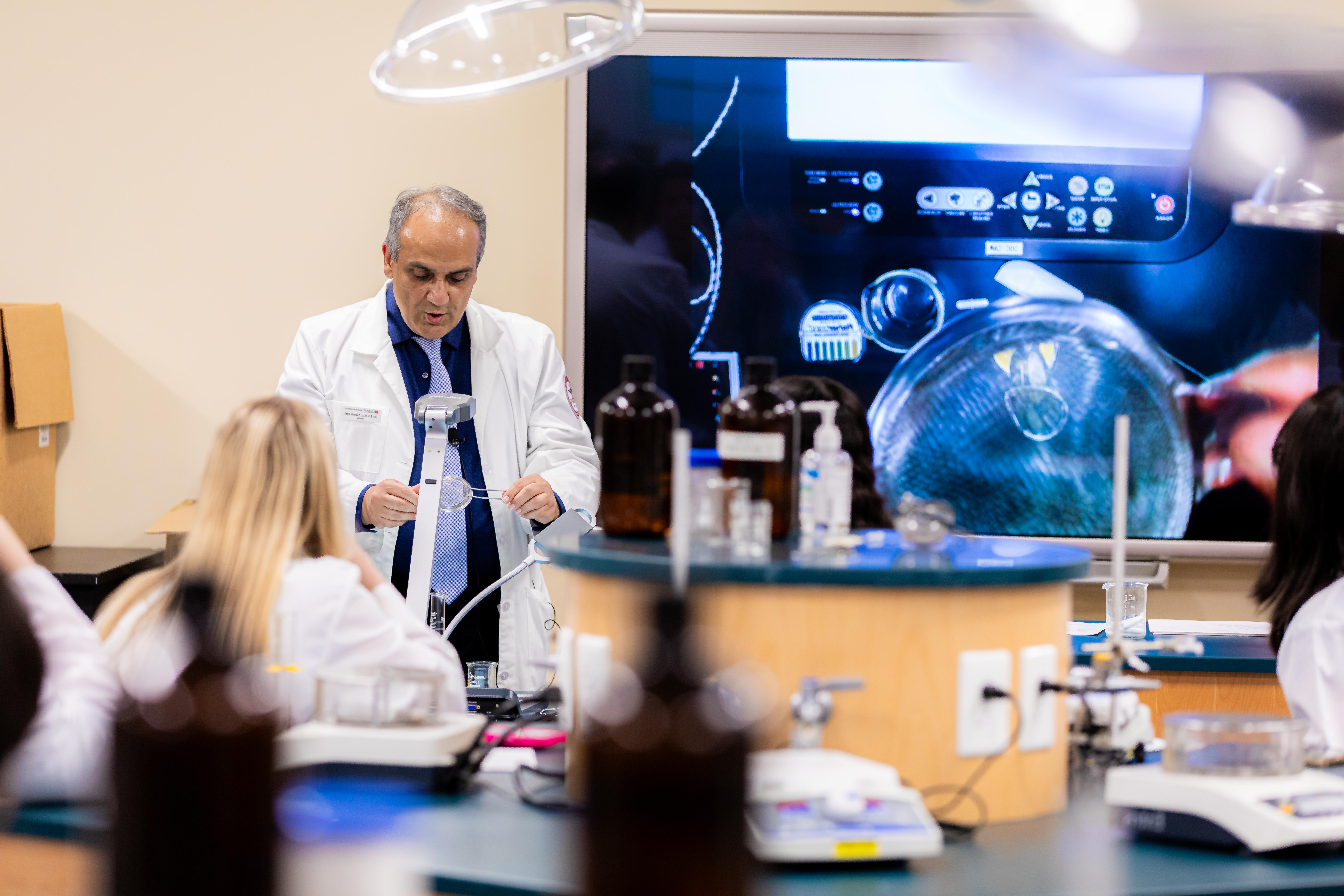 A man lecturing in the front of a lab with a lab coat on